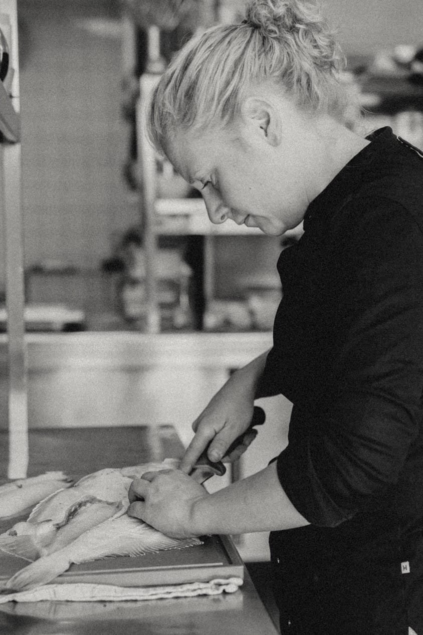 Een vrouw in een zwarte chef-kok outfit snijdt vis op een aanrecht, met focus op haar handelingen in een keukenomgeving.