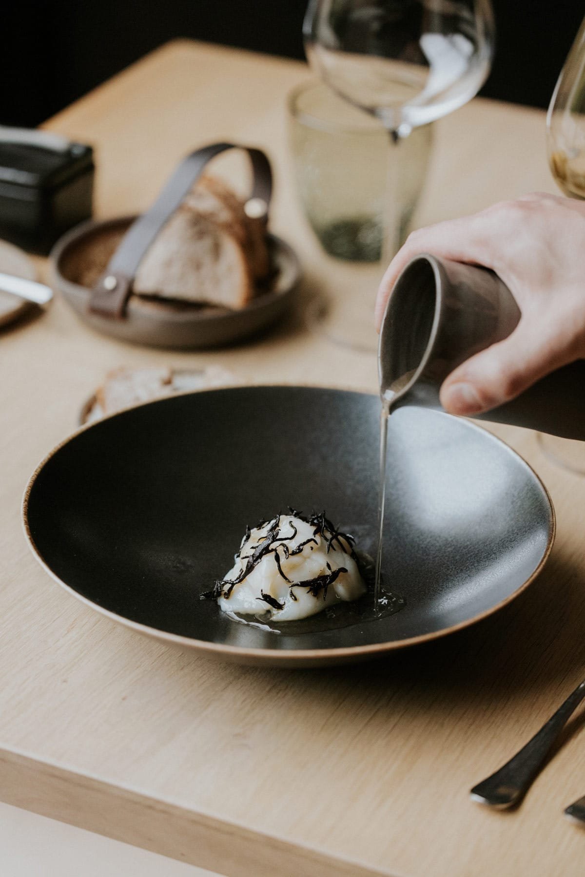 Een hand giet vloeistof uit een zwarte kan over een gerecht met een romige textuur en zwarte garnering op een zwart bord, voorzien van een houten tafel met een glazen wijn en een mandje met brood op de achtergrond.