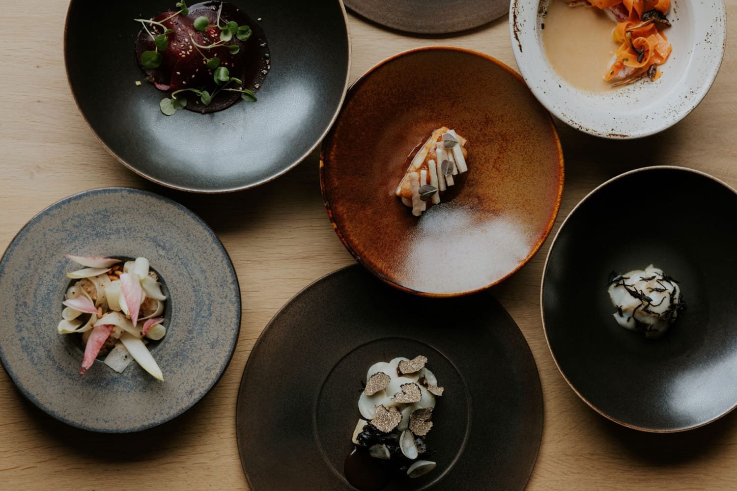 Een overhead beeld van verschillende elegante borden met creatief gepresenteerde gerechten, waaronder kleurrijke ingredi&euml;nten en garnituren, geserveerd op een houten tafel.