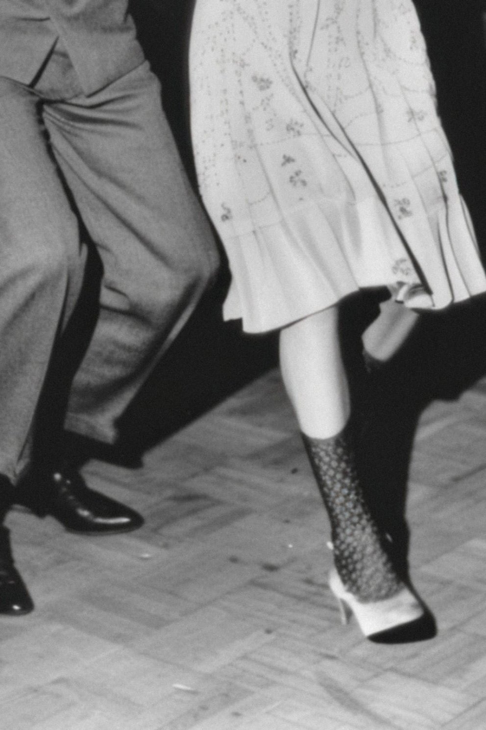 Beweging van dansende benen, met een man in een grijze broek en een vrouw in een lichte jurk met hoge hakken en een netpanty.