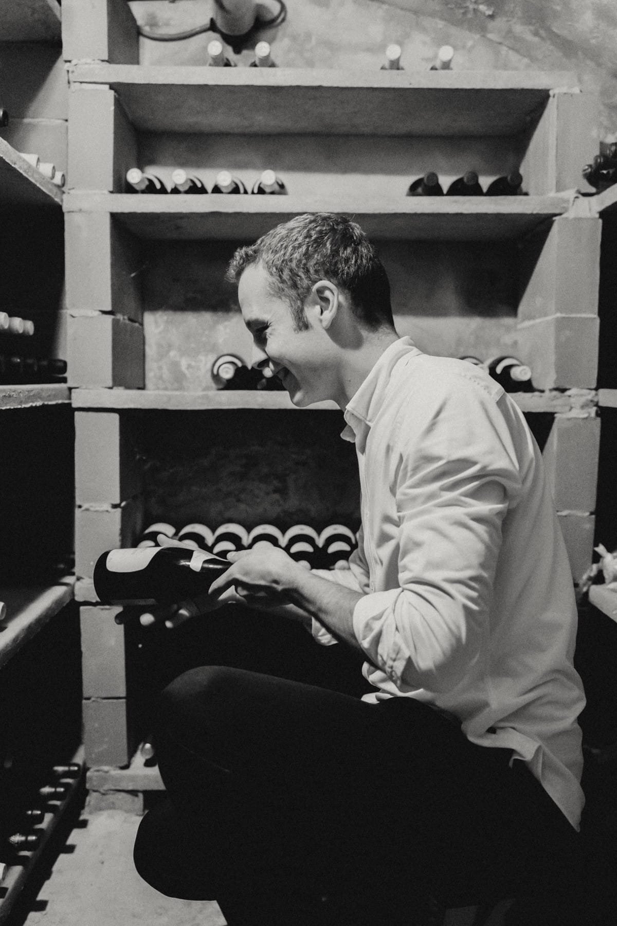 Een man in een wit shirt kijkt glimlachend naar een fles wijn in een wijnkelder, omringd door rijen met wijnflessen.